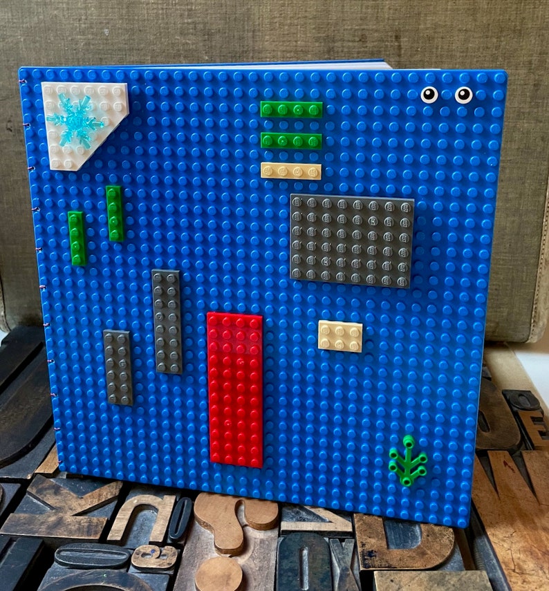 Large coptic bound blank book made from blue LEGO baseplates that measure 10 inches square. The book is decorated with 25 LEGO pieces in various colors. The paper is 70 pound paper suitable for drawing. The book is shown standing from the front.