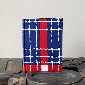 May include: A hardcover journal with a vibrant geometric pattern. The cover features a grid design with blue squares, white lines, and a central red stripe. The journal is standing upright on a dark surface.