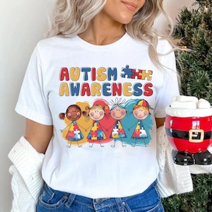 K&ouml;nnte beinhalten: Wei&szlig;es T-Shirt mit dem Text "AUTISM AWARENESS" und einer Grafik von vier Cartoon-Kindern, die sich an den H&auml;nden halten. Jedes Kind tr&auml;gt ein T-Shirt mit einem Puzzle-Motiv.