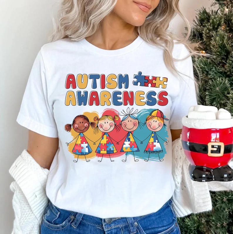 K&ouml;nnte beinhalten: Wei&szlig;es T-Shirt mit dem Text "AUTISM AWARENESS" und einer Grafik von vier Cartoon-Kindern, die sich an den H&auml;nden halten. Jedes Kind tr&auml;gt ein T-Shirt mit einem Puzzle-Motiv.