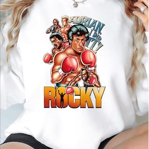 Op de afbeelding: Witte trui met een grafische print van een bokscene met Sylvester Stallone als Rocky Balboa. De grafiek bevat de tekst "Adrian I Did It!" en "Rocky".