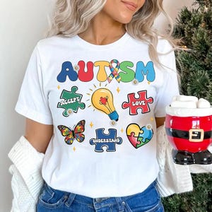 K&ouml;nnte beinhalten: Wei&szlig;es T-Shirt mit dem Wort "AUTISMUS" in Regenbogenfarben und Puzzle-Teilen mit den Worten "Akzeptieren", "Liebe" und "Verstehen".