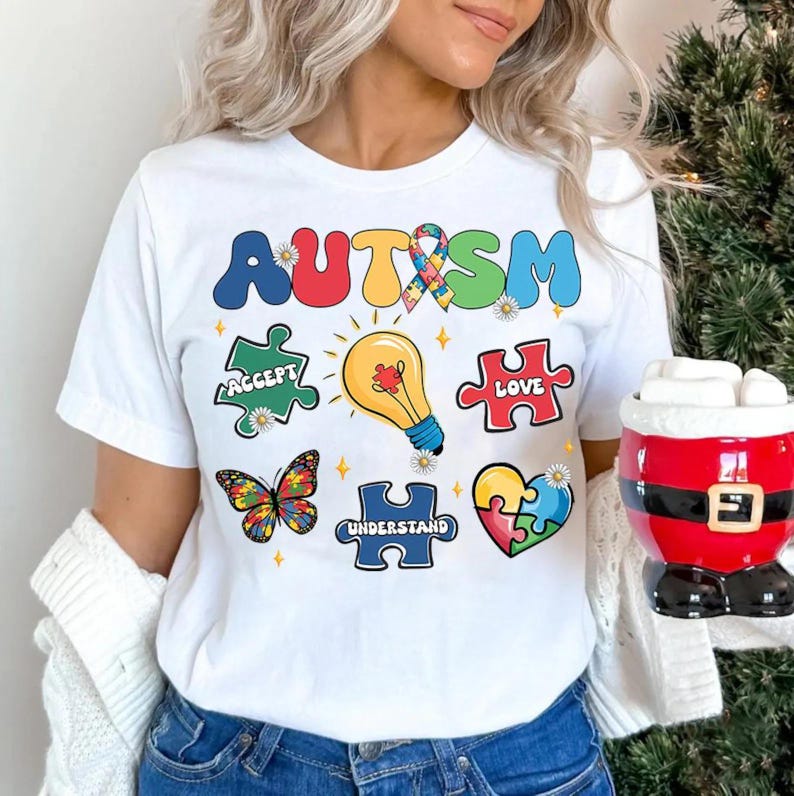 K&ouml;nnte beinhalten: Wei&szlig;es T-Shirt mit dem Wort "AUTISMUS" in Regenbogenfarben und Puzzle-Teilen mit den Worten "Akzeptieren", "Liebe" und "Verstehen".