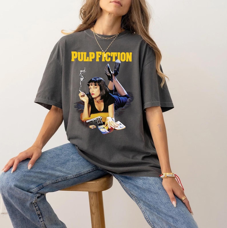 Op de afbeelding: Een antracietgrijs T-shirt met een grafische print van Mia Wallace uit de film Pulp Fiction. De tekst "PULP FICTION" is in geel boven de print gedrukt.