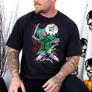 Op de afbeelding: Zwart T-shirt met een afbeelding van Jason Voorhees uit de film "Vrijdag de 13e Deel 3" die een machete vasthoudt en een hockeymasker draagt. De tekst "Vrijdag de 13e Deel 3" staat op het shirt gedrukt.