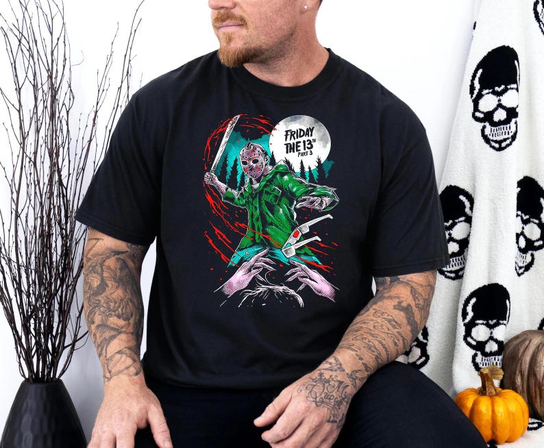 Op de afbeelding: Zwart T-shirt met een afbeelding van Jason Voorhees uit de film "Vrijdag de 13e Deel 3" die een machete vasthoudt en een hockeymasker draagt. De tekst "Vrijdag de 13e Deel 3" staat op het shirt gedrukt.