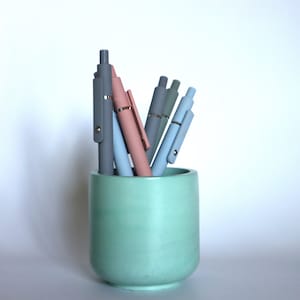 May include: A light green ceramic pencil holder with a variety of colored pens inside. The pens are gray, light blue, and pink.