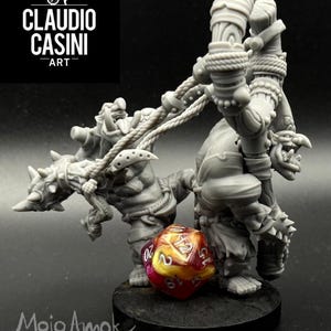 May include: A gray miniature figure of a fantasy creature with a weapon, standing on a black base. A multi-colored, twenty-sided die rests in front of the figure. The text "CLAUDIO CASINI ART" and "Mojo Amok miniatures" are visible.