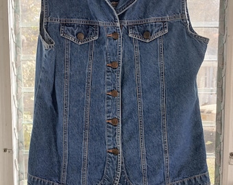 May include: A blue denim vest with a collared neckline and button-down closure. The vest has two front pockets and a curved hemline. It is hanging on a black hanger in front of a window.