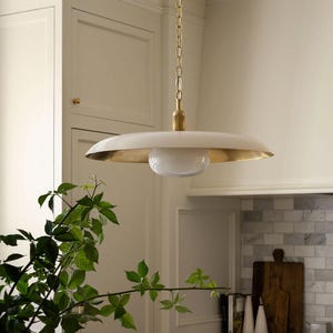 May include: A pendant light fixture featuring a wide, shallow, off-white metal shade with a metallic gold interior finish. A central, spherical white glass globe is suspended beneath the shade, connected by a brass-toned rod to a metal chain.