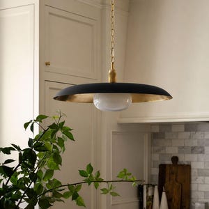 May include: A pendant light fixture featuring a wide, matte black saucer-shaped shade with a metallic gold interior finish. A central white glass globe bulb is suspended from a brass-toned metal rod and a matching chain link ceiling mount.