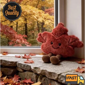May include: A plush toy shaped like a maple leaf with a deep red, fuzzy texture, featuring a small embroidered smiling face and two brown corduroy legs. The leaf is positioned on a stone ledge near a window with autumn foliage visible.
