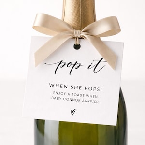May include: A champagne bottle with a gold foil top and a cream-colored ribbon tied around the neck. A white tag with the words "pop it" and other text is attached to the bottle. The text reads "WHEN SHE POPS! ENJOY A TOAST WHEN BABY CONNOR ARRIVES" with a heart symbol.