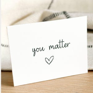 May include: A white greeting card with the words "you matter" in a handwritten font, and a small heart below the text. The card is on a wooden surface, with a soft, textured background. The Max & Bella logo is at the bottom.