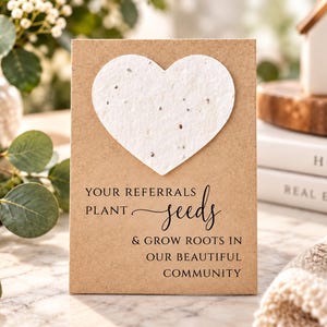 May include: A rectangular kraft paper card featuring a large, textured white heart made of seed paper at the top. The card displays the black printed text: YOUR REFERRALS PLANT seeds & GROW ROOTS IN OUR BEAUTIFUL COMMUNITY in a mix of fonts.