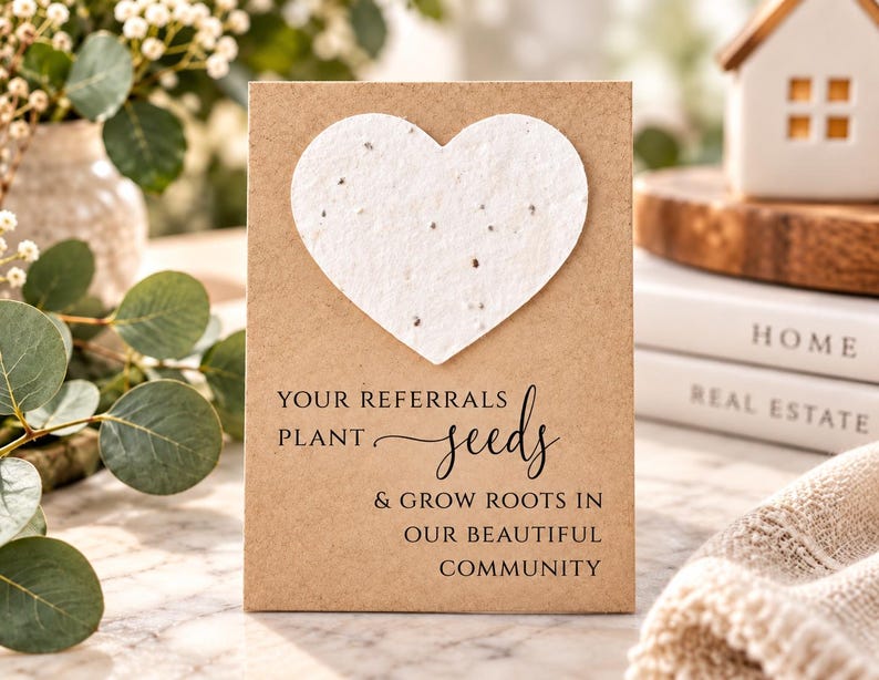May include: A rectangular kraft paper card featuring a large, textured white heart made of seed paper at the top. The card displays the black printed text: YOUR REFERRALS PLANT seeds & GROW ROOTS IN OUR BEAUTIFUL COMMUNITY in a mix of fonts.