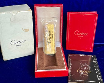 May include: A gold Cartier lighter in a red box with a white box and a black booklet. The box has the Cartier logo and the words "Paris" on it.