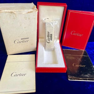May include: A collection of Cartier items, including a silver lighter in a red box, a cream-colored box labeled "Briquiet Cartier", and several booklets. The lighter is marked "Cartier PARIS".