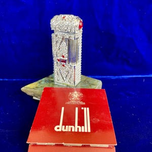 May include: A silver-toned lighter with red accents and a decorative design, resting on a stone base. The lighter is accompanied by a red box with the Dunhill logo and the text "Lighter Flints". The background is a deep blue.