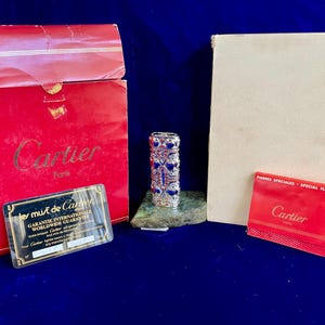 May include: A Cartier lighter with a silver and blue design, presented with its original red box and warranty card. The box is red with gold lettering. The lighter is on a green stone base.
