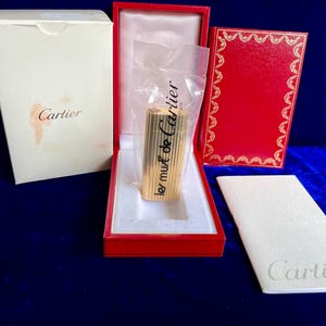 May include: A gold-toned Cartier lighter with vertical ribbed detailing, encased in a clear plastic sleeve. It is presented in a red velvet-lined box with a white outer sleeve, a red decorative box lid, and a white Cartier branded booklet.
