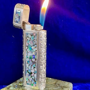 Könnte beinhalten: Ein silbernes Feuerzeug mit offenem Deckel und brennender Flamme. Das Feuerzeug ist mit einem Mosaik aus schillernden Steinen verziert. Der Körper des Feuerzeugs hat ein strukturiertes Muster. Die Flamme ist gelb und blau.