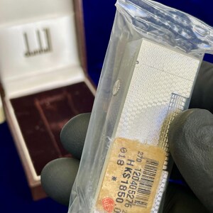May include: A silver Dunhill lighter in a clear plastic bag, with a textured wave pattern. The lighter has a rectangular shape and a small sticker with text and a barcode. A brown box with the Dunhill logo is in the background.