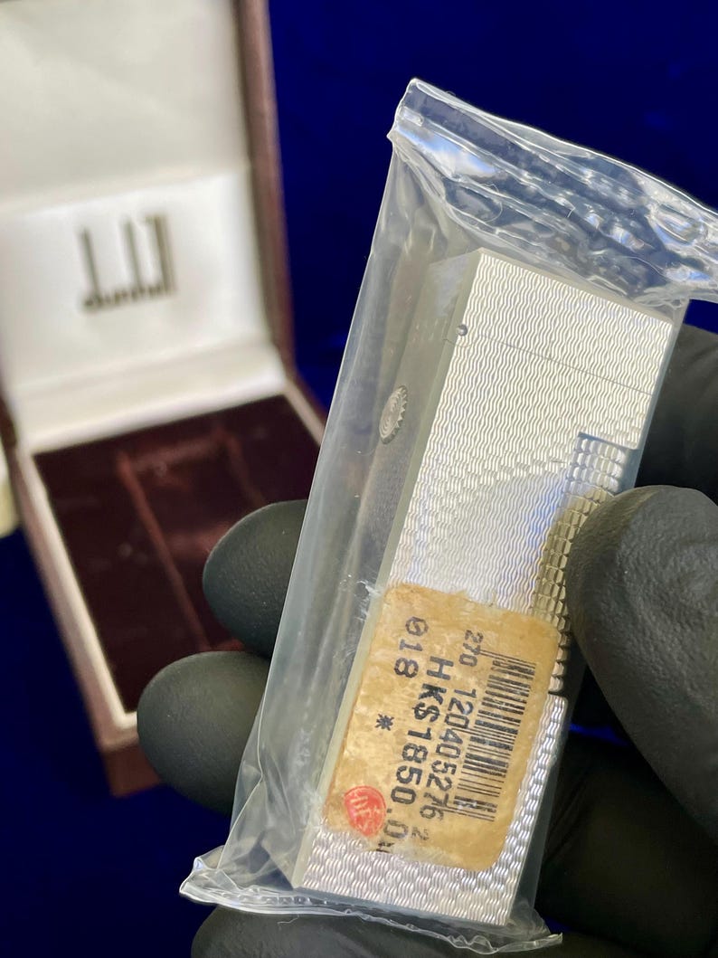 May include: A silver Dunhill lighter in a clear plastic bag, with a textured wave pattern. The lighter has a rectangular shape and a small sticker with text and a barcode. A brown box with the Dunhill logo is in the background.