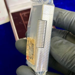 May include: A silver-colored lighter with a textured pattern, still in its original plastic packaging. The lighter has a rectangular shape with a smaller, textured section. A price tag is visible.