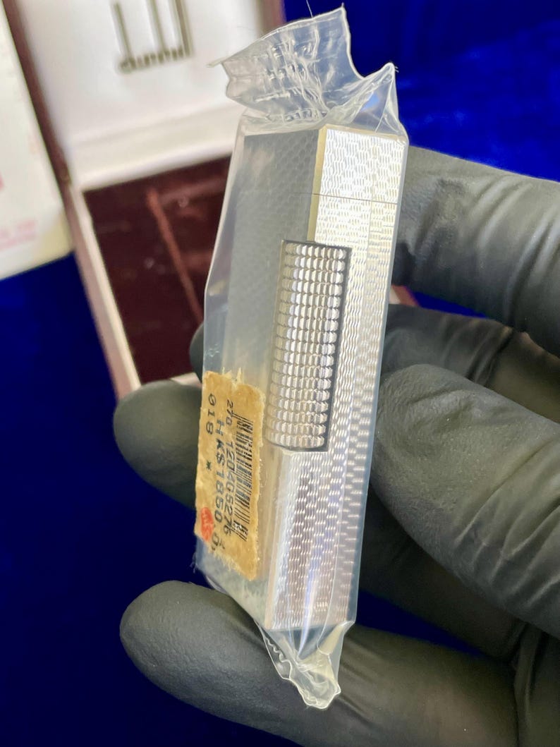 May include: A silver-colored lighter with a textured pattern, still in its original plastic packaging. The lighter has a rectangular shape with a smaller, textured section. A price tag is visible.