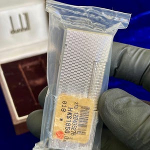 May include: A silver Dunhill lighter in a plastic bag, with a textured pattern. The lighter is rectangular with a ridged edge. A price tag is attached. The Dunhill logo is visible on the box.
