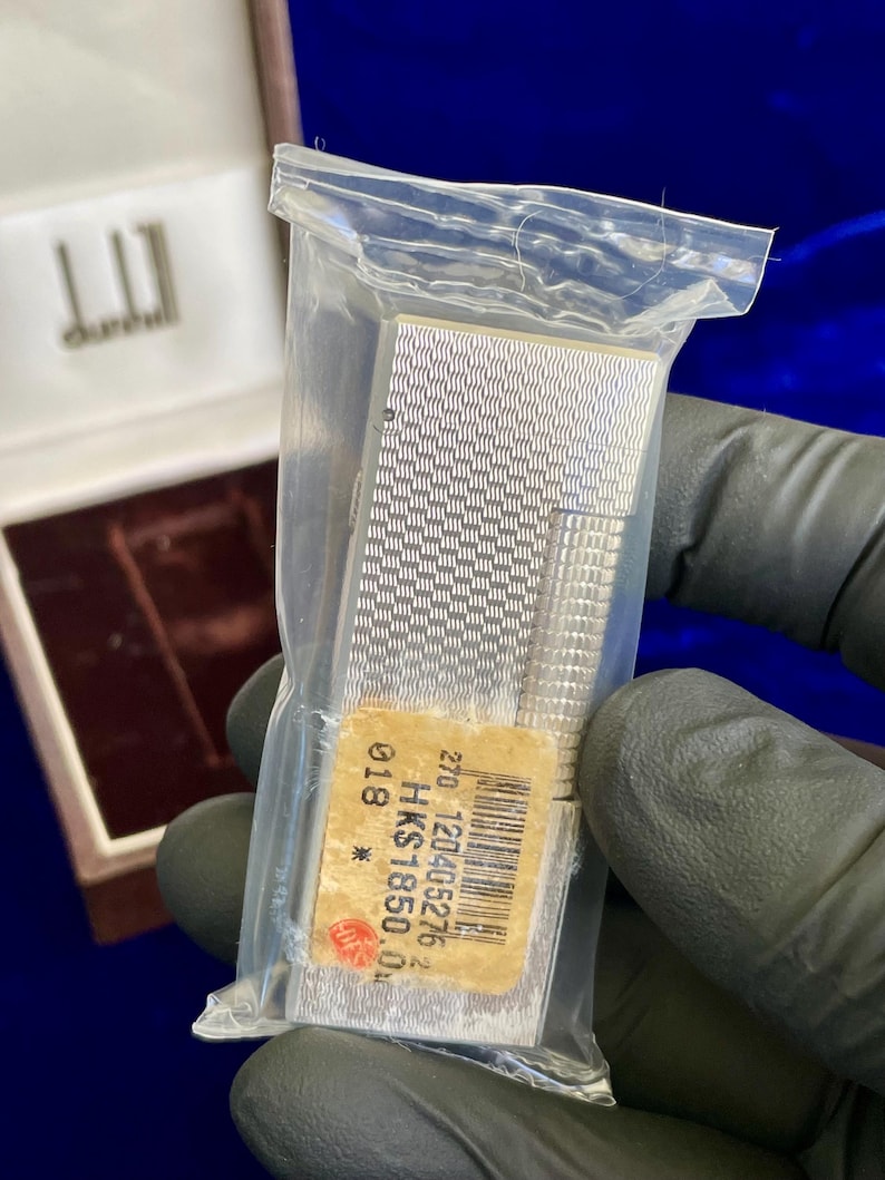 May include: A silver Dunhill lighter in a plastic bag, with a textured pattern. The lighter is rectangular with a ridged edge. A price tag is attached. The Dunhill logo is visible on the box.