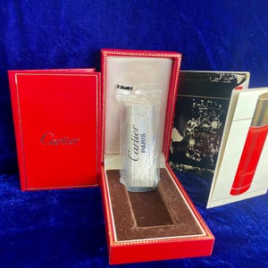 May include: A silver-colored Cartier perfume bottle with the words "Cartier PARIS" is displayed in a red box. The box is open, revealing the bottle inside. A red box with the Cartier logo and a perfume box are also visible.