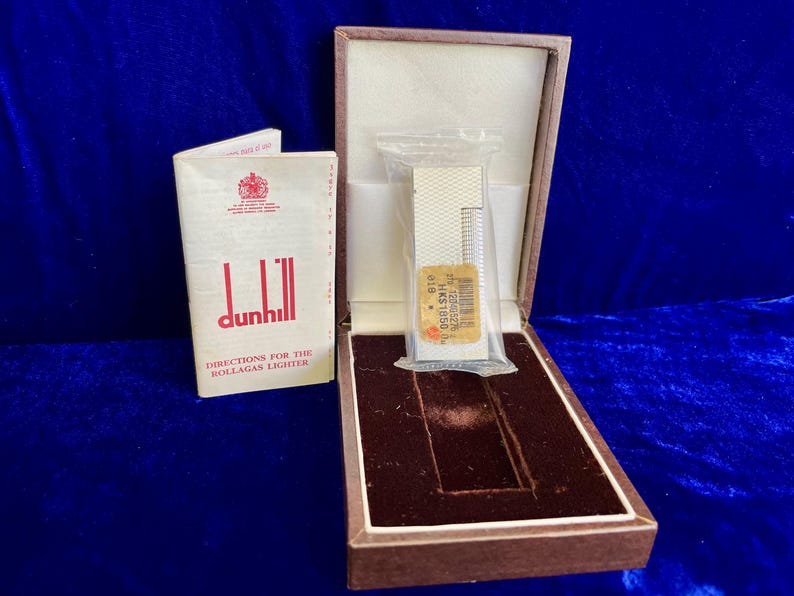 May include: A vintage Dunhill Rollagas lighter in its original box, with the instruction booklet. The lighter is silver-toned and comes with a clear plastic protective cover. The box is brown with a velvet interior.