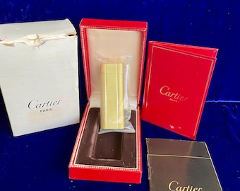 Puede incluir: Un encendedor Cartier dorado en una caja roja con un interior de terciopelo marrón. La caja tiene un borde dorado y el logotipo de Cartier está impreso en la parte superior. El encendedor está en una bolsa de plástico y la caja está sobre una superficie azul.