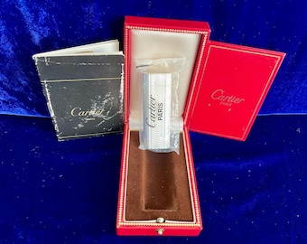 May include: A silver Cartier lighter in a red box with the Cartier logo. The lighter is in a plastic sleeve and has the words "Cartier PARIS" printed on it. A black box and a red box are also visible.
