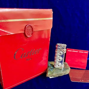 May include: A red Cartier box with gold trim and a seal, next to a decorative lighter with blue and silver accents. A red card and a silver card are also present. The box has the Cartier logo in gold.