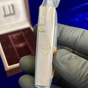 May include: A silver Dunhill cigarette case, encased in clear plastic. The case has a textured, patterned surface and a circular button. The Dunhill logo is visible on the open box in the background. The case is rectangular and appears to be new.
