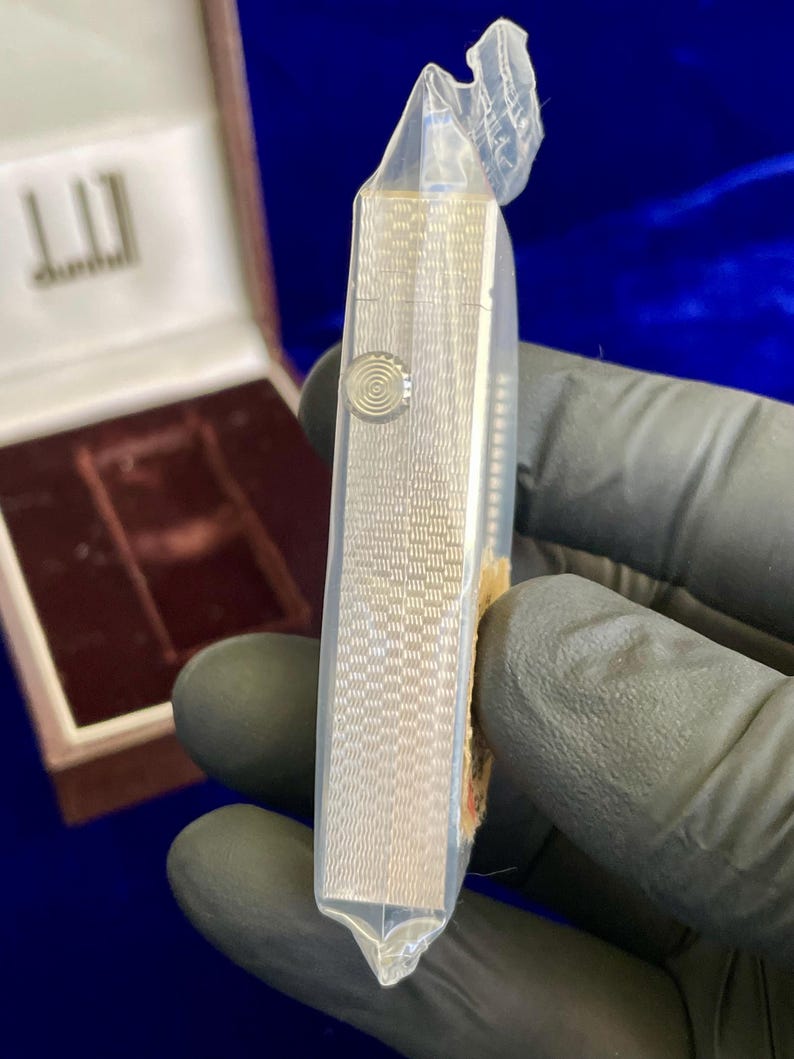 May include: A silver Dunhill cigarette case, encased in clear plastic. The case has a textured, patterned surface and a circular button. The Dunhill logo is visible on the open box in the background. The case is rectangular and appears to be new.