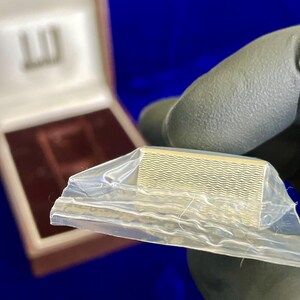 May include: A gold-colored, rectangular money clip with a wavy pattern, partially encased in clear plastic. The clip is held by a gloved hand, with a box in the background. The box has a logo on the lid.