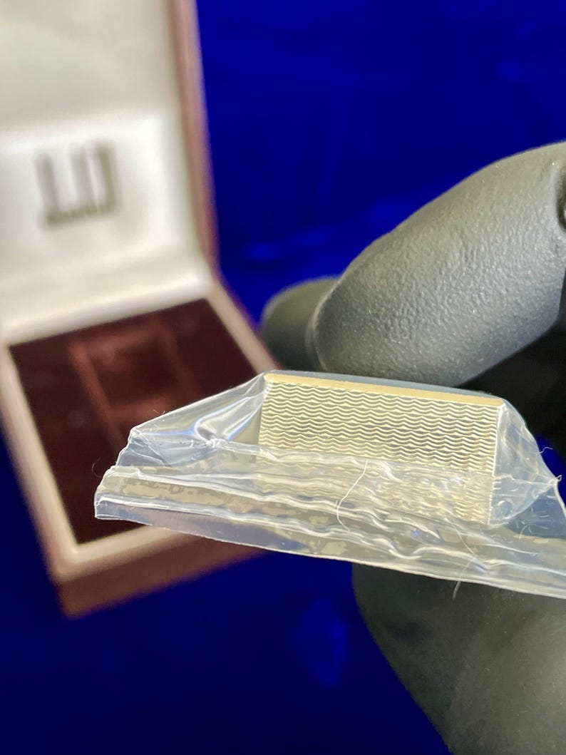 May include: A gold-colored, rectangular money clip with a wavy pattern, partially encased in clear plastic. The clip is held by a gloved hand, with a box in the background. The box has a logo on the lid.