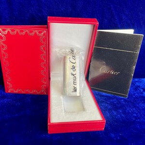 May include: A silver-colored object with the text "les must de Cartier" is displayed in a red box with a white interior. A red box with gold trim and a black box with the Cartier logo are also present. The items are on a blue surface.