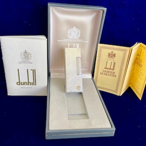 May include: A silver Dunhill dress lighter in its original presentation box, accompanied by a booklet and a guarantee card. The lighter has a ribbed design and a gold-colored accent. The box is gray with a light pink interior.