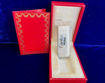 May include: A silver-colored Cartier Paris lighter in a red box. The lighter is rectangular with a textured surface and the Cartier logo. The red box has a gold-colored decorative border and a white interior.