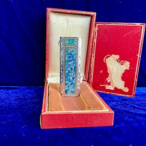 Könnte beinhalten: Ein silbernes Vintage-Feuerzeug mit einem Mosaik aus blauen und türkisfarbenen Steinen, präsentiert in einer roten Samtbox. Die Box ist geöffnet und zeigt das Feuerzeug und sein plüschiges Inneres. Die rote Box hat eine goldene Umrandung.