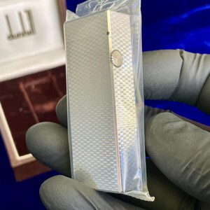 May include: A silver-colored Dunhill lighter with a textured, checkered pattern, encased in a clear plastic bag. The lighter has a circular design on the side and is rectangular in shape. The Dunhill logo is visible in the background.