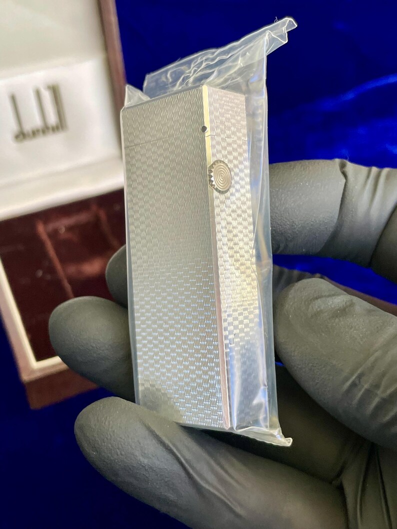 May include: A silver-colored Dunhill lighter with a textured, checkered pattern, encased in a clear plastic bag. The lighter has a circular design on the side and is rectangular in shape. The Dunhill logo is visible in the background.