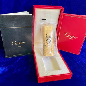 May include: A gold-colored Cartier lighter in a clear plastic sleeve, presented in a red box with a white interior. The box is open, revealing the lighter and a black booklet. The Cartier logo is visible on the box and booklet.