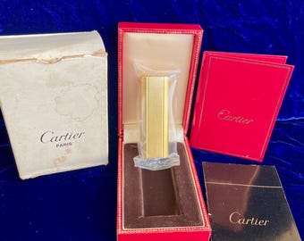 May include: A gold Cartier lighter in its red box with a white box and a booklet. The lighter is in a clear plastic bag. The box has the Cartier logo and the words "Paris" on it.