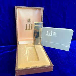 May include: A vintage Dunhill lighter in its original presentation box. The lighter has a gold finish with a tortoiseshell pattern. The box is brown with a tan interior and the Dunhill logo is visible. A booklet labeled "Lighters" is also present.